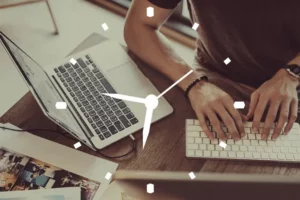 يدان تعملان على كمبيوتر مع ساعة، تمثل إدارة الوقت في التعليم عن بعد وتأثيرها على الأداء الأكاديمي.